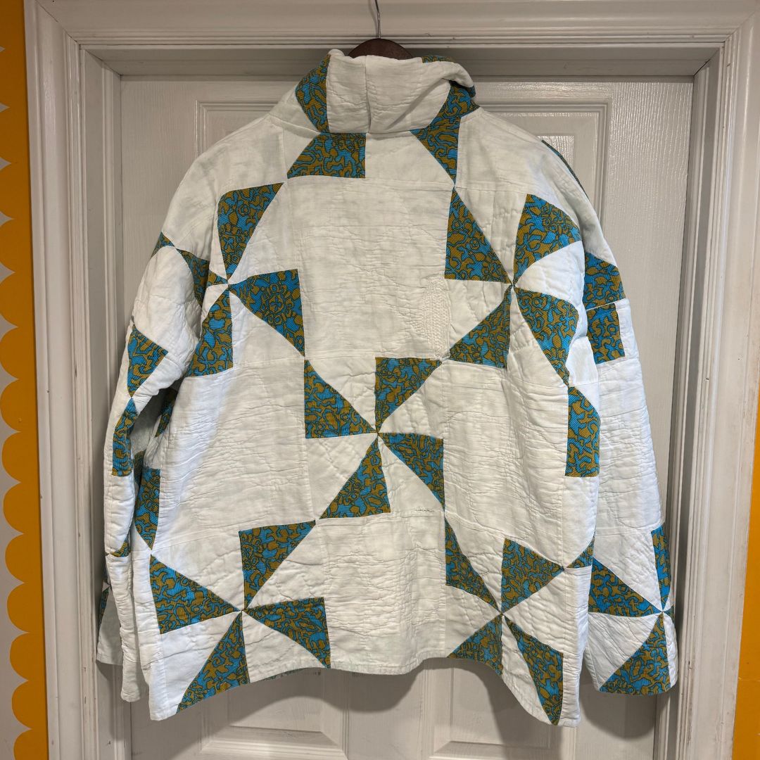 A quilted jacket with a high mock neck and hood, featuring a white base with geometric triangles patterned in teal and yellow, hanging on a wooden hanger in front of a white paneled door.