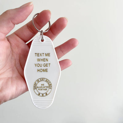 A hand holds a white, motel-style key tag with a silver keyring. The tag has gold text that says 