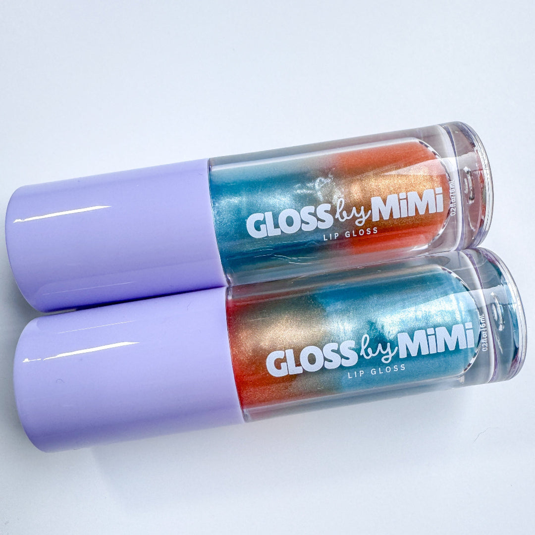 Two tubes of GLOSS by MIMI The Life of a Showgirl Lip Gloss with lavender caps and multicoloured shimmer gloss in blue, silver, and orange gradients, styled to evoke a vibrant, theatrical aesthetic.