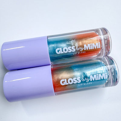 Two tubes of GLOSS by MIMI The Life of a Showgirl Lip Gloss with lavender caps and multicoloured shimmer gloss in blue, silver, and orange gradients, styled to evoke a vibrant, theatrical aesthetic.
