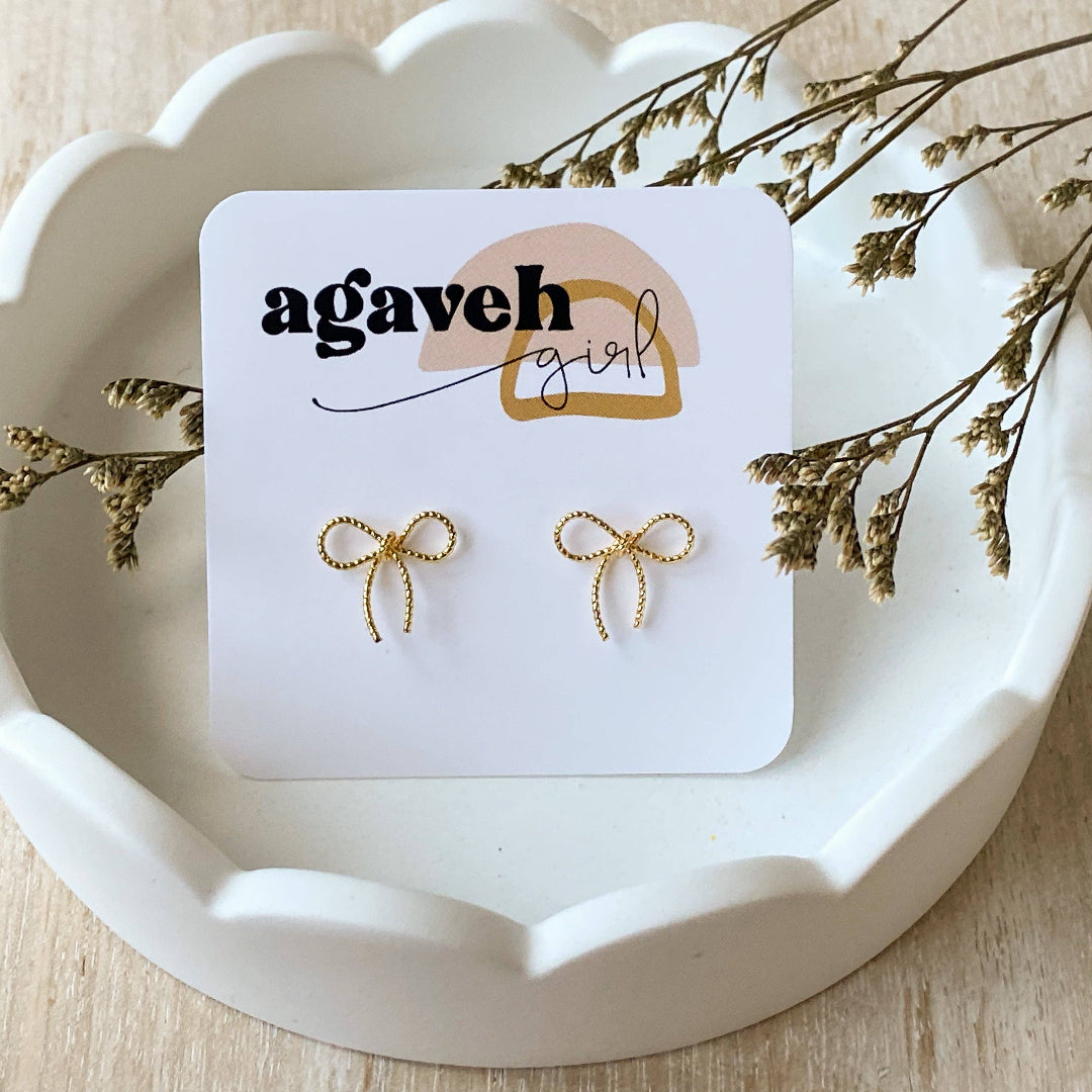 A pair of Agaveh Girl Elle Bow Studs, featuring a minimalist, gold wire bow design, displayed on a branded card with the text 'agaveh girl,' against a scalloped dish background with dried floral accents. The earrings are dainty and delicate, available in Ottawa, Ontario, Canada.