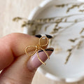 A pair of Agaveh Girl Elle Bow Studs, featuring a minimalist, gold wire bow design, displayed on a branded card with the text 'agaveh girl,' against a scalloped dish background with dried floral accents. The earrings are dainty and delicate, available in Ottawa, Ontario, Canada.