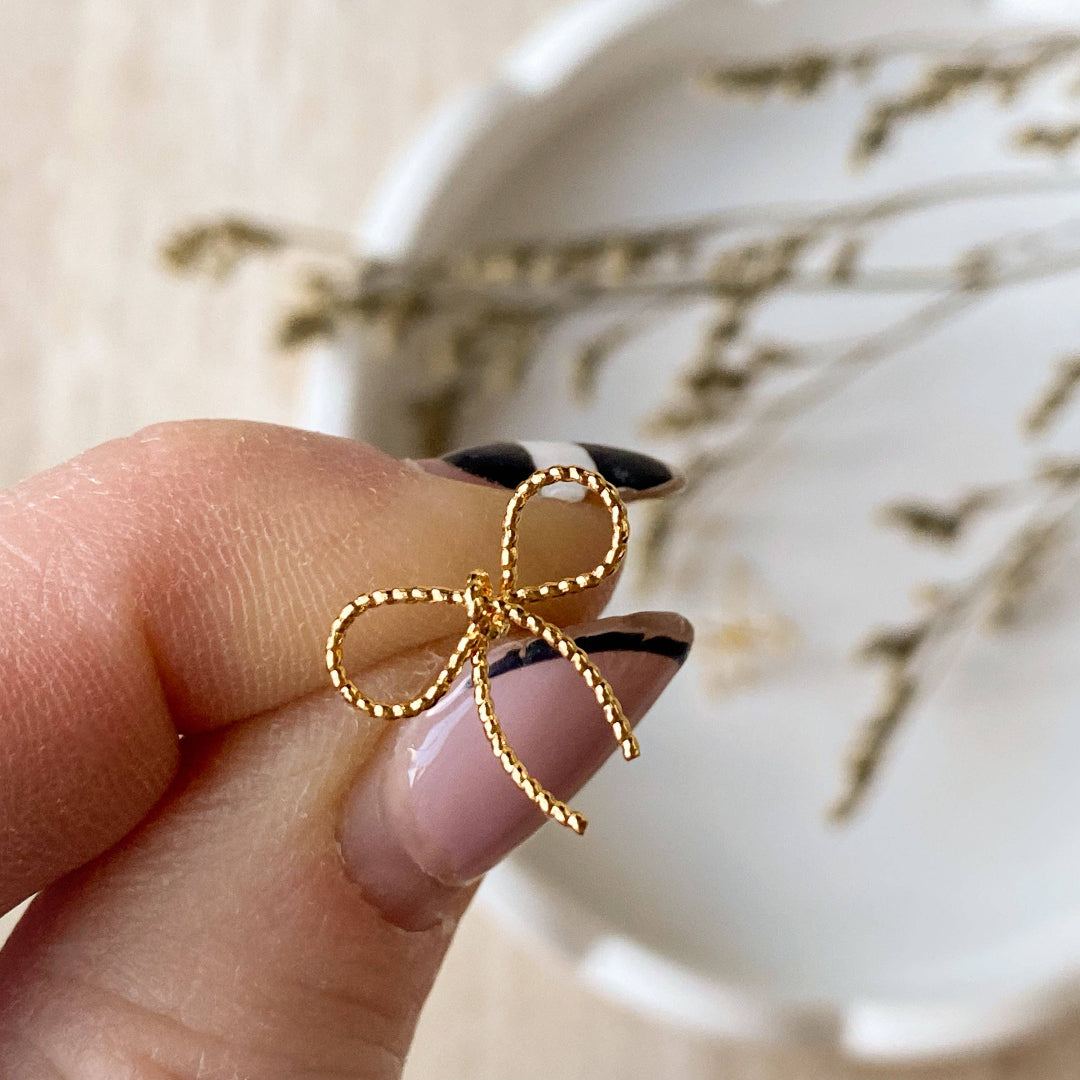 A pair of Agaveh Girl Elle Bow Studs, featuring a minimalist, gold wire bow design, displayed on a branded card with the text 'agaveh girl,' against a scalloped dish background with dried floral accents. The earrings are dainty and delicate, available in Ottawa, Ontario, Canada.
