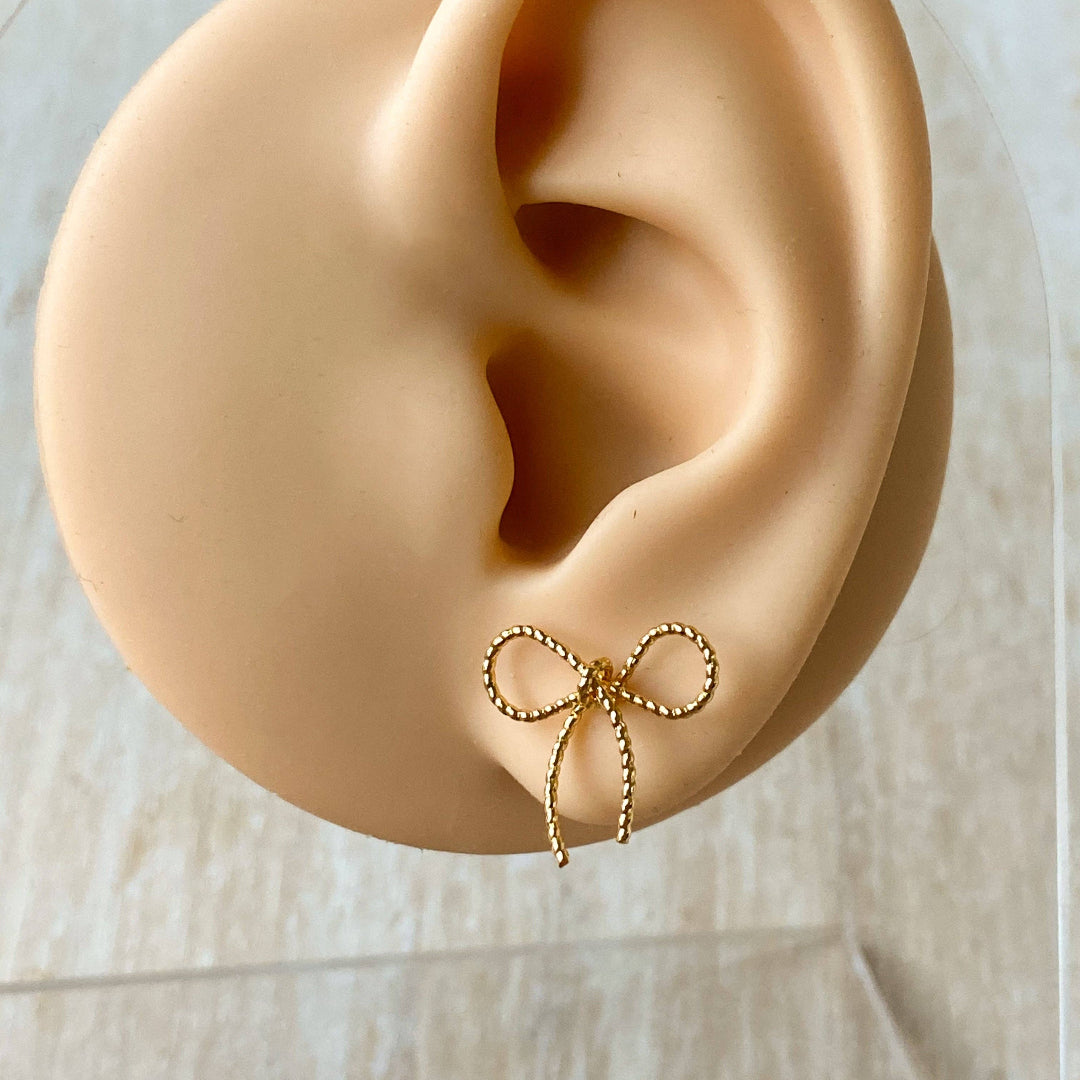 A pair of Agaveh Girl Elle Bow Studs, featuring a minimalist, gold wire bow design, displayed on a branded card with the text 'agaveh girl,' against a scalloped dish background with dried floral accents. The earrings are dainty and delicate, available in Ottawa, Ontario, Canada.