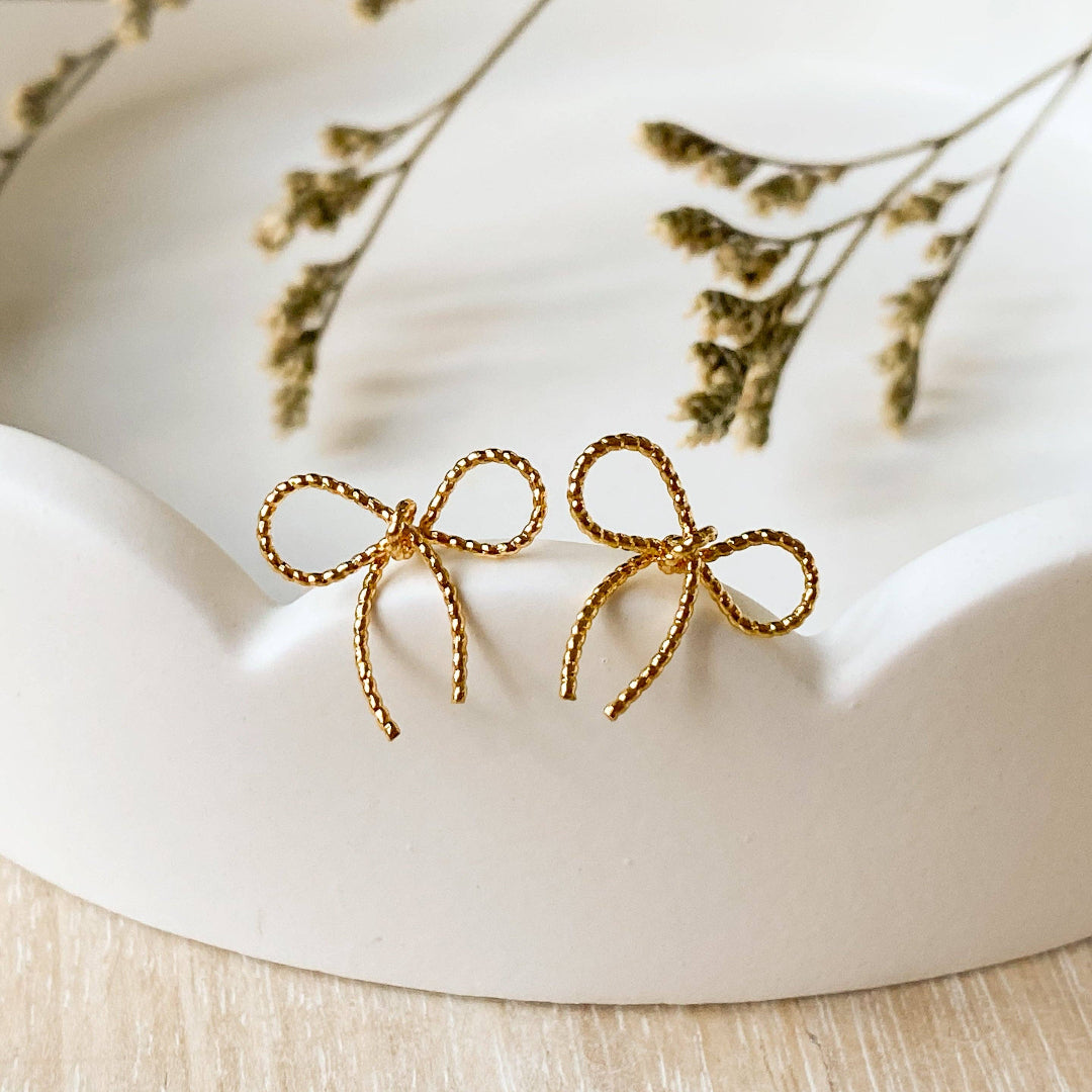 A pair of Agaveh Girl Elle Bow Studs, featuring a minimalist, gold wire bow design, displayed on a branded card with the text 'agaveh girl,' against a scalloped dish background with dried floral accents. The earrings are dainty and delicate, available in Ottawa, Ontario, Canada.