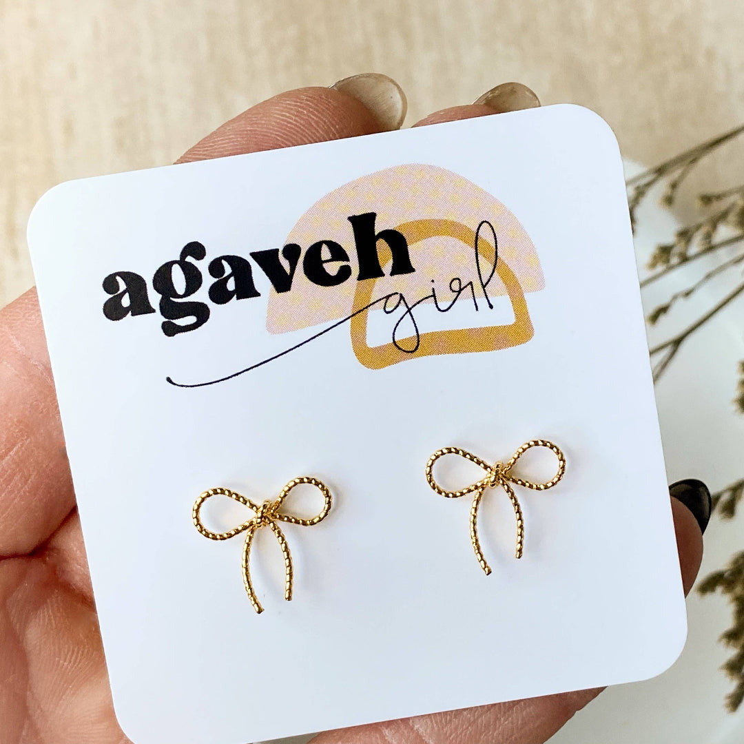 A pair of Agaveh Girl Elle Bow Studs, featuring a minimalist, gold wire bow design, displayed on a branded card with the text 'agaveh girl,' against a scalloped dish background with dried floral accents. The earrings are dainty and delicate, available in Ottawa, Ontario, Canada.
