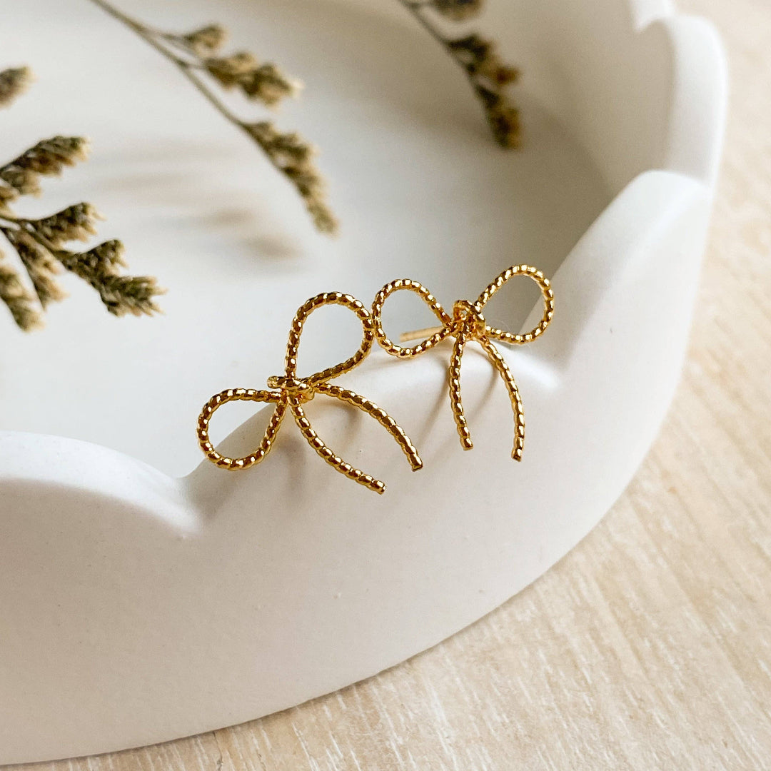 A pair of Agaveh Girl Elle Bow Studs, featuring a minimalist, gold wire bow design, displayed on a branded card with the text 'agaveh girl,' against a scalloped dish background with dried floral accents. The earrings are dainty and delicate, available in Ottawa, Ontario, Canada.