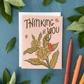 Greeting card with floral design and 'Thinking of You' text on a blue background with green leaves and pens.