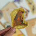 A hand holds a die-cut sticker featuring a brown bear sitting thoughtfully with the text 'TIME FOR A PONDER' on a warm mustard-yellow background. The scene is softly blurred with other colorful stickers in the background.