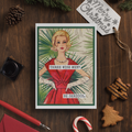 Vintage-style Christmas card with a woman in a red dress on a wooden surface with holiday decorations.