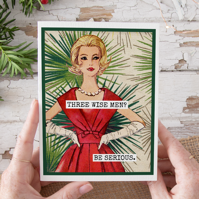 Card with a woman in a red dress and text 'Three Wise Men? Be Serious.' held by hands against a rustic background.