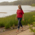 A woman walking barefoot on a sandy path next to a grassy shoreline with a calm lake and forest in the background, wearing a red short-sleeve top and dark cuffed jeans.