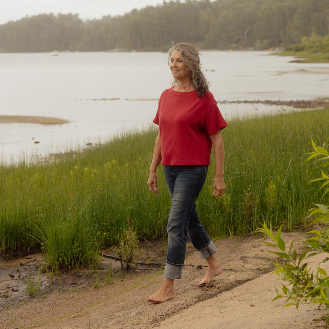 A woman walking barefoot on a sandy path next to a grassy shoreline with a calm lake and forest in the background, wearing a red short-sleeve top and dark cuffed jeans.
