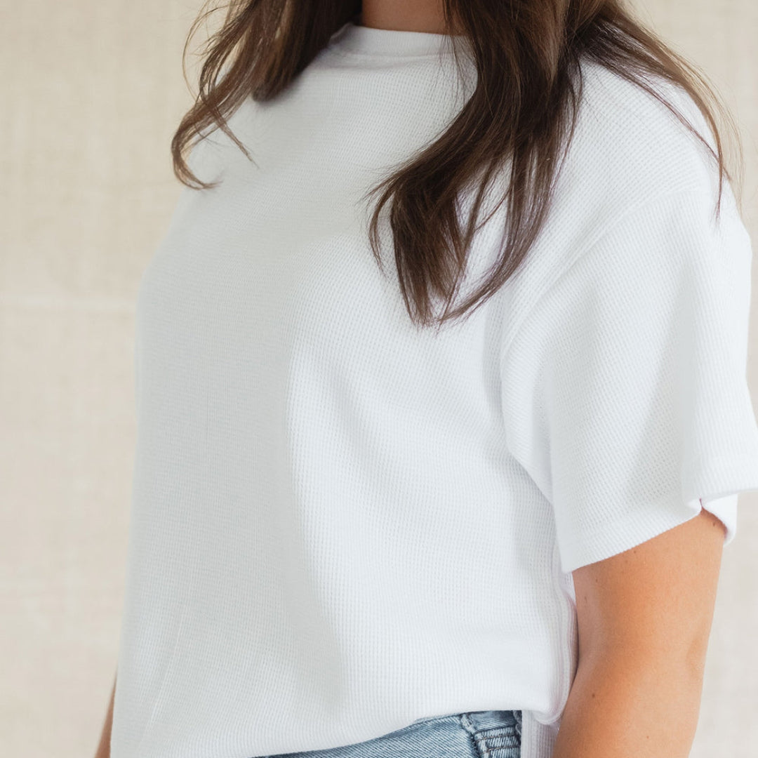 A close-up of a woman wearing a white short-sleeve ribbed knit top with a side-slit hem, paired with light-wash blue jeans against a neutral beige background.