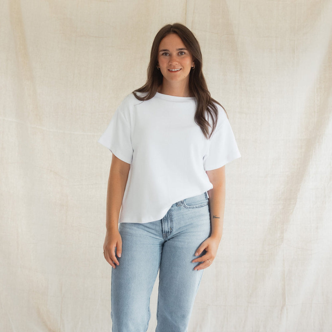 A woman stands barefoot against a draped beige fabric backdrop, wearing a loose white short-sleeve top and light blue straight-leg jeans.