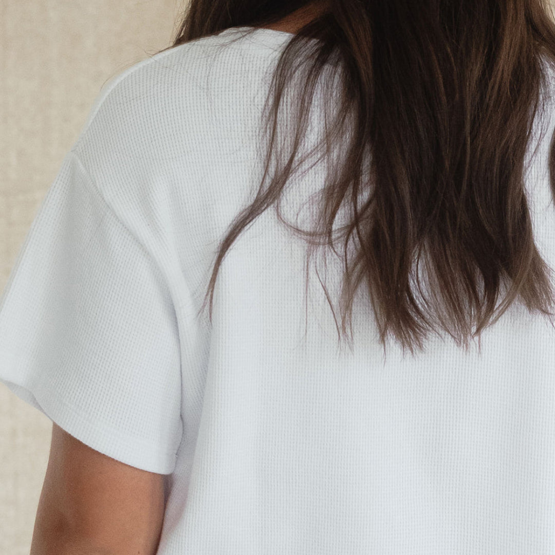 A close-up back view of a person with long dark brown hair wearing a white short-sleeve ribbed t-shirt against a neutral beige textured background.