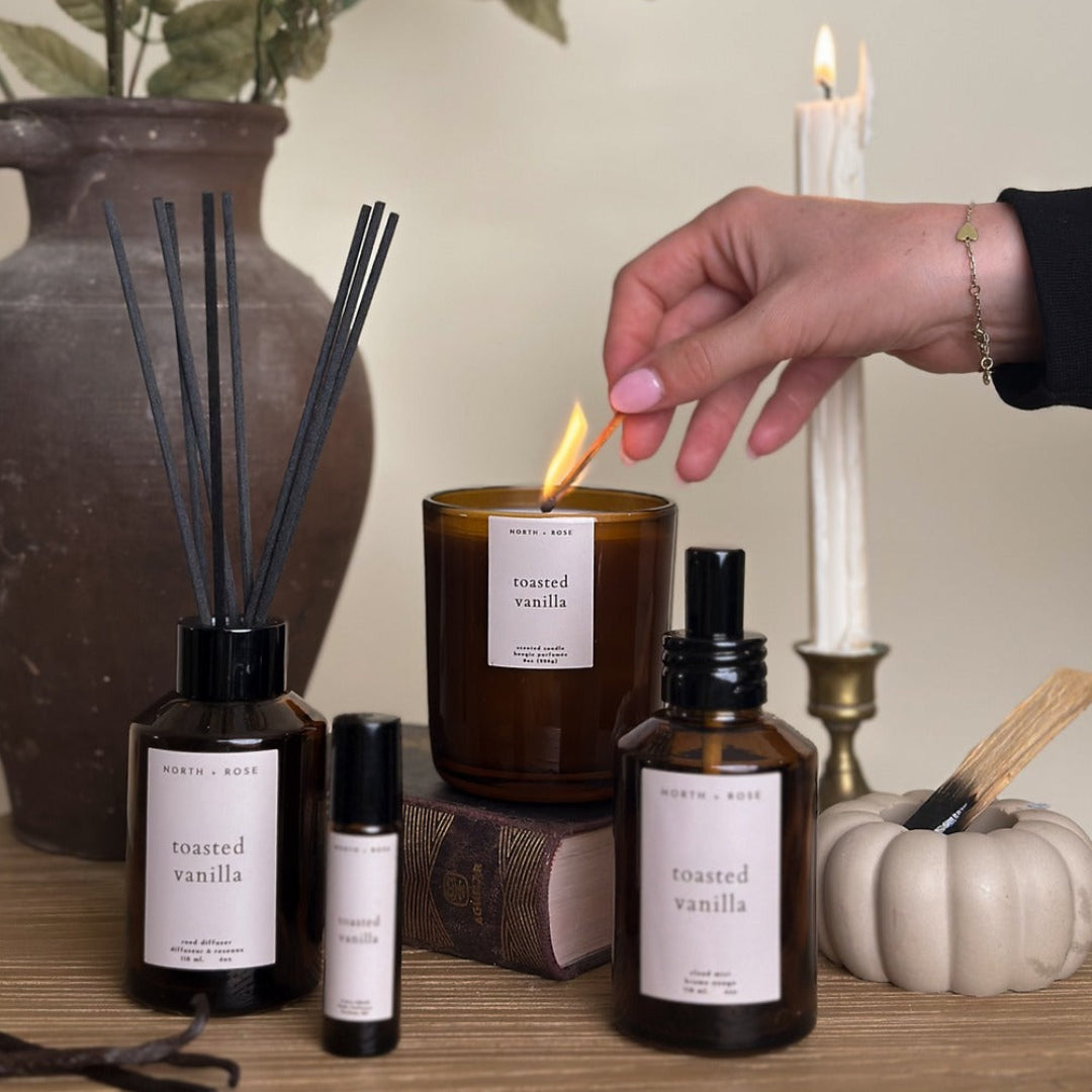Person lighting a candle labeled 'toasted vanilla' surrounded by diffusers and bottles on a wooden surface.