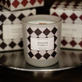 A soy candle in a frosted glass jar labeled 'TOASTED VANILLA' sits on a reflective metal tray, with matching patterned product boxes behind it in a dimly lit setting.