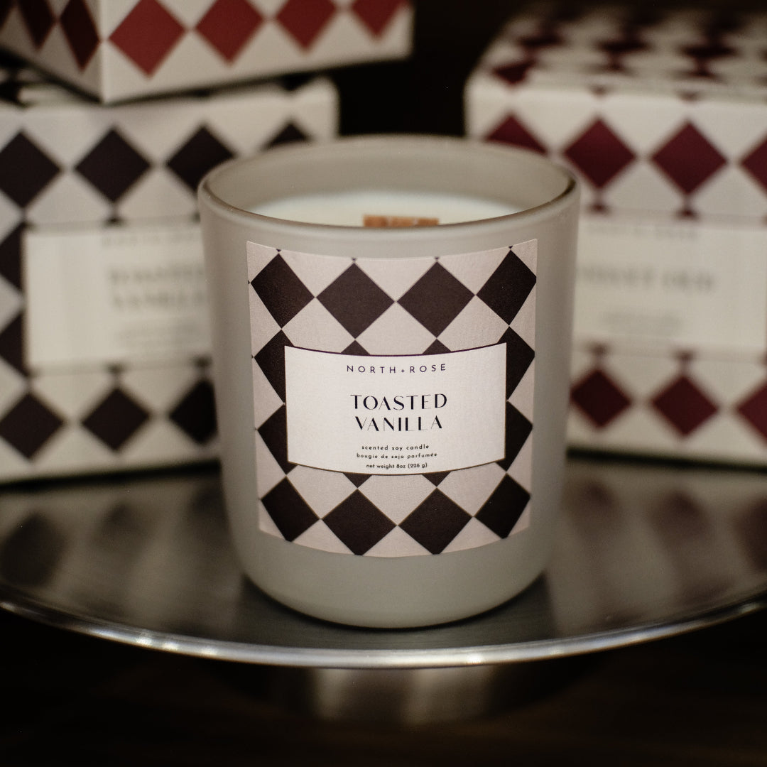 A soy candle in a frosted glass jar labeled 'TOASTED VANILLA' sits on a reflective metal tray, with matching patterned product boxes behind it in a dimly lit setting.