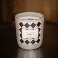 A North & Rose 'Toasted Vanilla' scented candle in a white glass jar with a black and white diamond pattern label, resting on a dark wooden surface against a dark wood background.