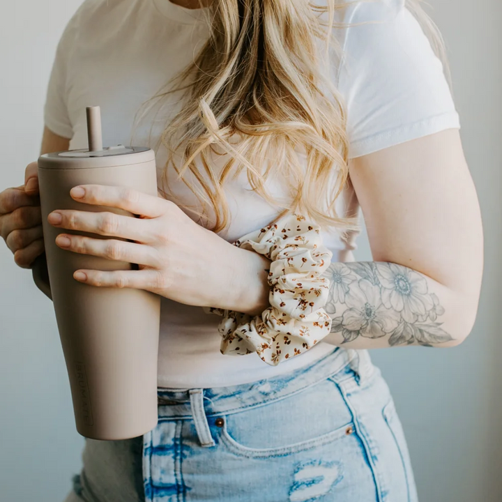 A cream-colored fabric scrunchie with a gathered, ruffled appearance. It features a delicate floral pattern in shades of brown, resembling dried flowers.