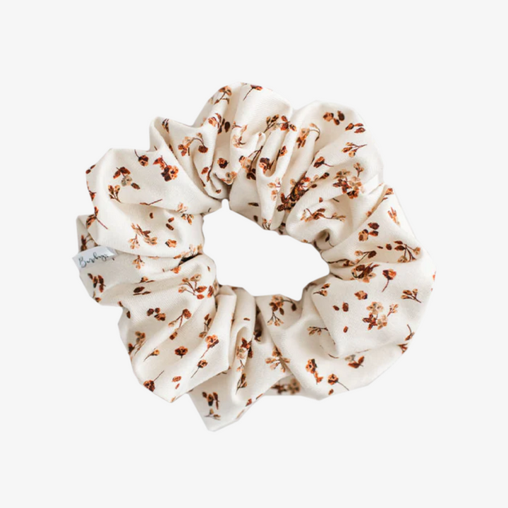 A cream-colored fabric scrunchie with a gathered, ruffled appearance. It features a delicate floral pattern in shades of brown, resembling dried flowers.