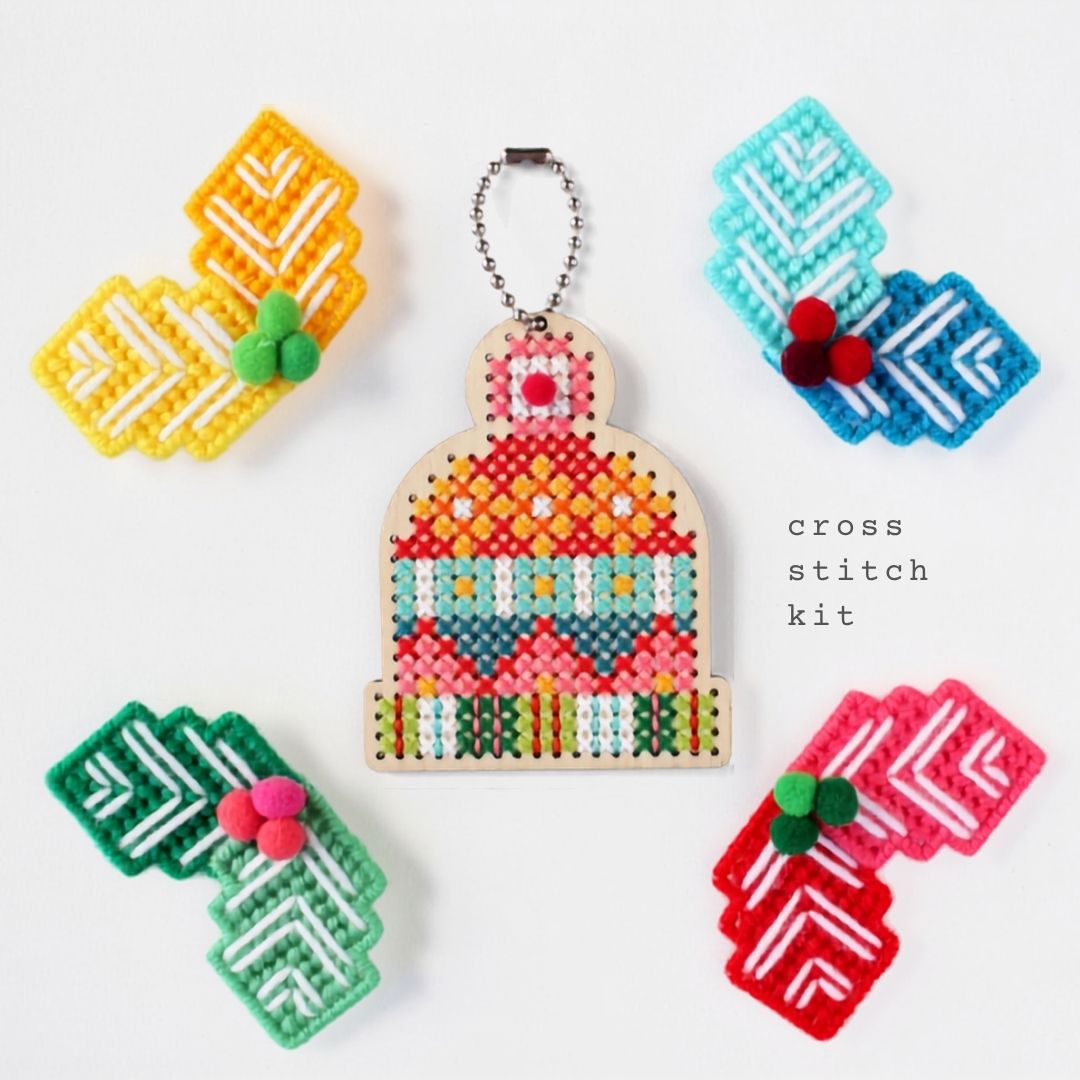 A cross stitch kit displayed on a white background, featuring a central completed bell-shaped cross stitch design with a hanging chain, surrounded by five colorful needlework starter pieces in yellow, blue, green, red, and pink, with the text 'cross stitch kit' on the right.