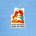 A die-cut sticker featuring an orange tabby kitten hugging a smaller kitten, with the text 'At least my trauma made me funny' below. The sticker is displayed on a textured light blue background.