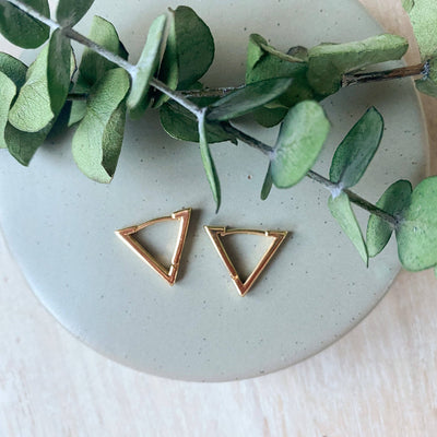 Gold triangle earrings on a light surface with green leaves.