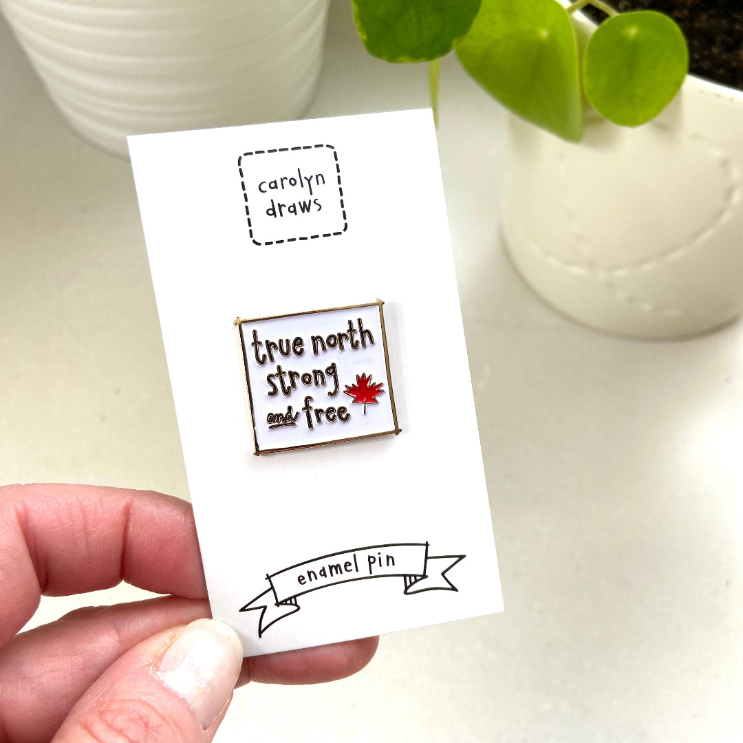 Enamel pin with 'true north strong and free' text and a maple leaf on a card held by a hand, with a plant in the background.