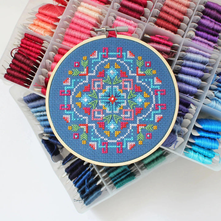 A completed cross-stitch project in a wooden hoop. The design features a colorful geometric pattern in shades of blue, red, yellow, and green. The project is displayed on a white surface with a small potted plant in the background. The text 