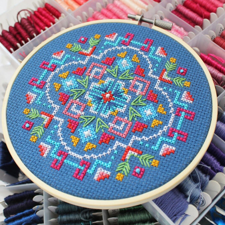 A completed cross-stitch project in a wooden hoop. The design features a colorful geometric pattern in shades of blue, red, yellow, and green. The project is displayed on a white surface with a small potted plant in the background. The text 
