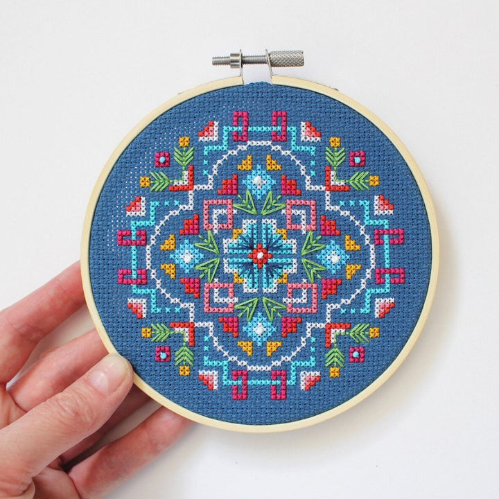 A completed cross-stitch project in a wooden hoop. The design features a colorful geometric pattern in shades of blue, red, yellow, and green. The project is displayed on a white surface with a small potted plant in the background. The text 