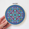 A completed cross-stitch project in a wooden hoop. The design features a colorful geometric pattern in shades of blue, red, yellow, and green. The project is displayed on a white surface with a small potted plant in the background. The text 