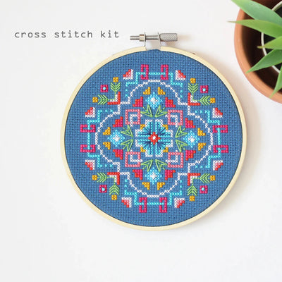 A completed cross-stitch project in a wooden hoop. The design features a colorful geometric pattern in shades of blue, red, yellow, and green. The project is displayed on a white surface with a small potted plant in the background. The text 