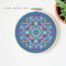 A completed cross-stitch project in a wooden hoop. The design features a colorful geometric pattern in shades of blue, red, yellow, and green. The project is displayed on a white surface with a small potted plant in the background. The text 