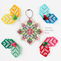 A cross stitch kit displayed on a white background, featuring a central wooden star-shaped sampler with a colorful geometric cross-stitch pattern, surrounded by five finished sample pieces in yellow, blue, green, pink, and red, along with the text 'cross stitch kit'.