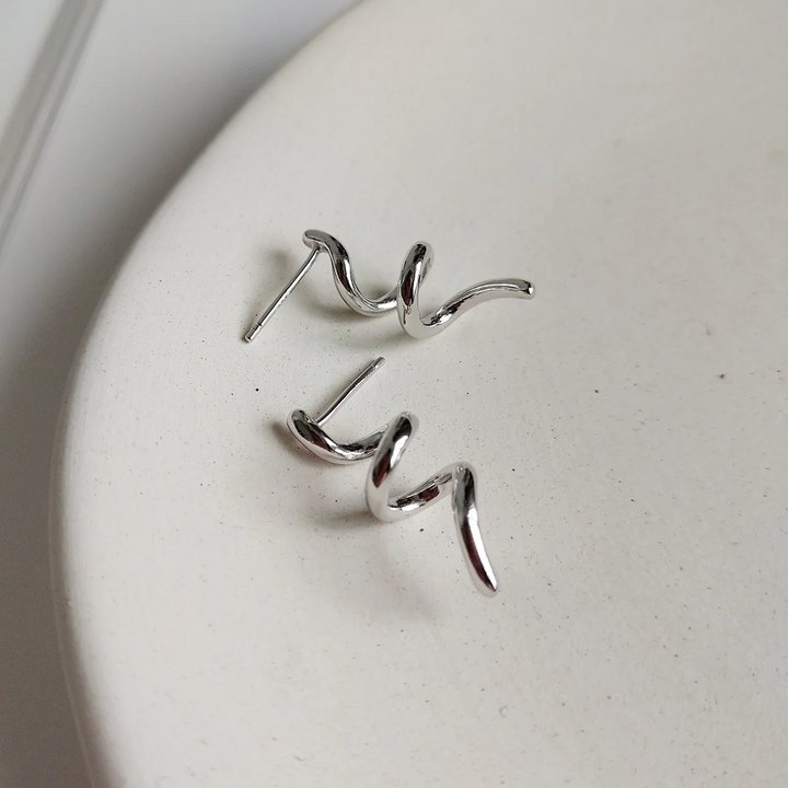 A pair of twisty coil earrings made of sterling silver, featuring a unique, abstract, and wavy design, displayed on a light-colored surface. These modern earrings are available in Ottawa, Ontario, Canada.