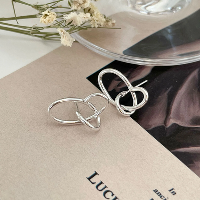 A pair of twisty knot stud earrings made of sterling silver, featuring a unique, abstract, and intertwined design, displayed on a book with dried flowers and a glass object. These modern earrings are available in Ottawa, Ontario, Canada.