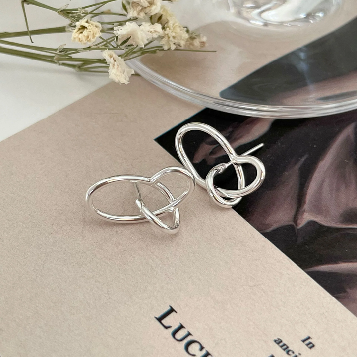 A pair of twisty knot stud earrings made of sterling silver, featuring a unique, abstract, and intertwined design, displayed on a book with dried flowers and a glass object. These modern earrings are available in Ottawa, Ontario, Canada.