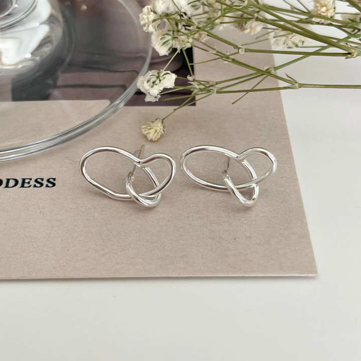 A pair of twisty knot stud earrings made of sterling silver, featuring a unique, abstract, and intertwined design, displayed on a book with dried flowers and a glass object. These modern earrings are available in Ottawa, Ontario, Canada.