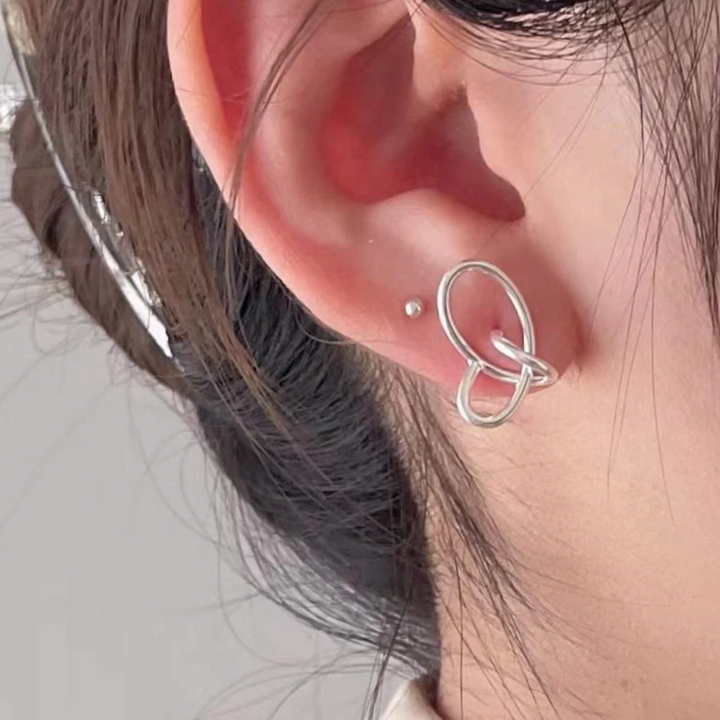 A pair of twisty knot stud earrings made of sterling silver, featuring a unique, abstract, and intertwined design, displayed on a book with dried flowers and a glass object. These modern earrings are available in Ottawa, Ontario, Canada.