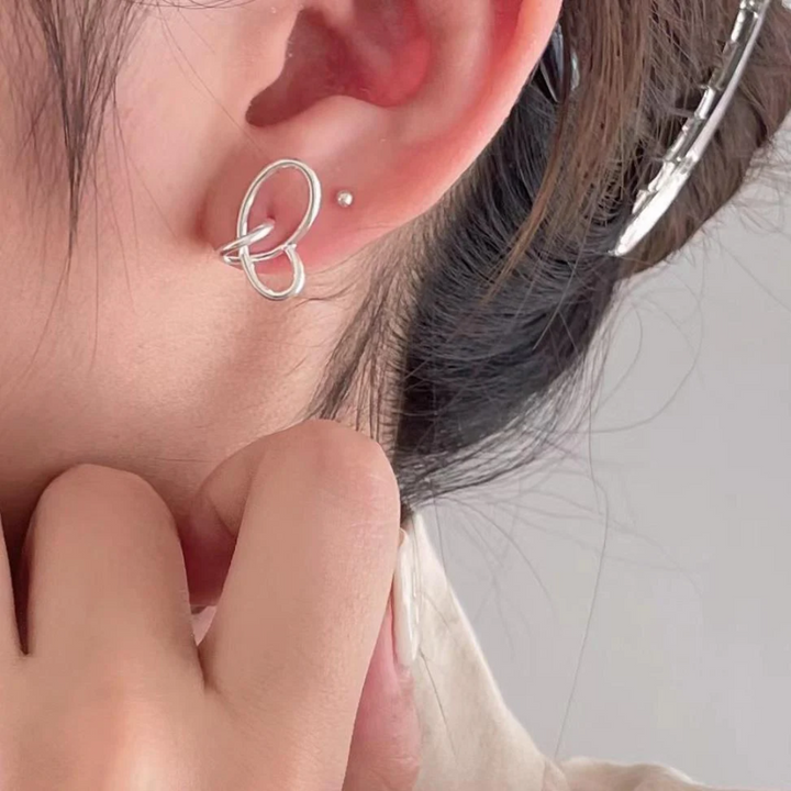 A pair of twisty knot stud earrings made of sterling silver, featuring a unique, abstract, and intertwined design, displayed on a book with dried flowers and a glass object. These modern earrings are available in Ottawa, Ontario, Canada.