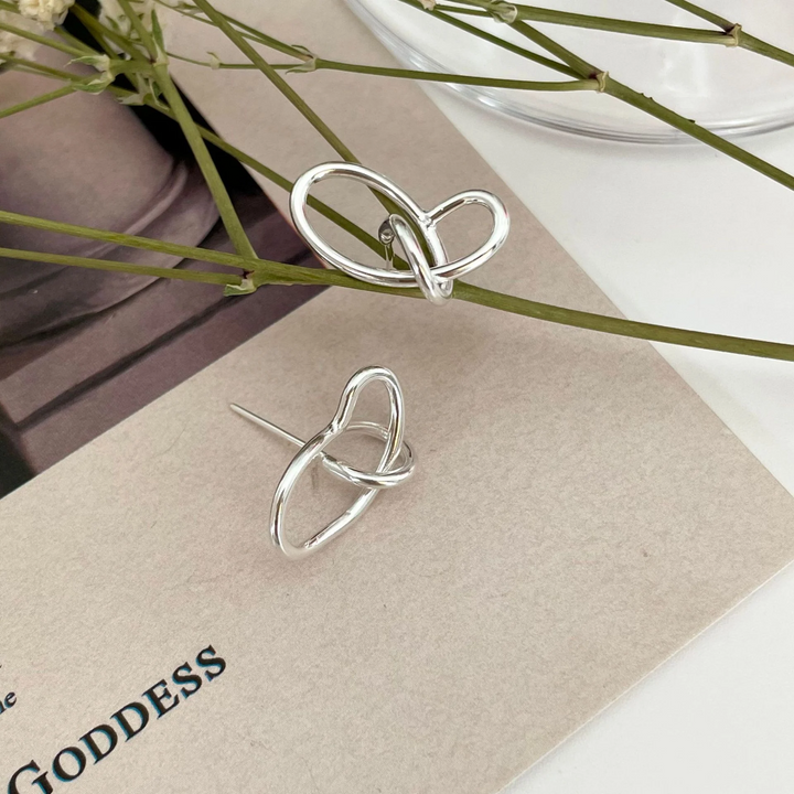 A pair of twisty knot stud earrings made of sterling silver, featuring a unique, abstract, and intertwined design, displayed on a book with dried flowers and a glass object. These modern earrings are available in Ottawa, Ontario, Canada.