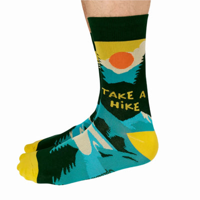 A pair of green socks with a hiking-themed design. The socks feature a mountain landscape, a setting sun, and the text 
