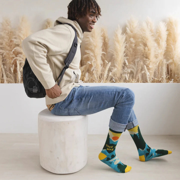 A pair of green socks with a hiking-themed design. The socks feature a mountain landscape, a setting sun, and the text 