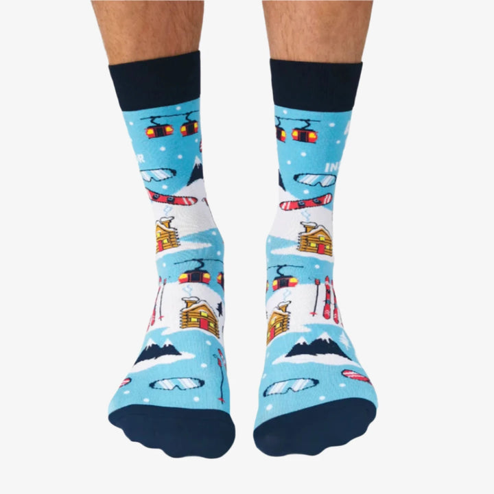 A pair of blue socks with a winter sports theme. The socks feature illustrations of skiers, snowboards, and a mountain cabin. The text 