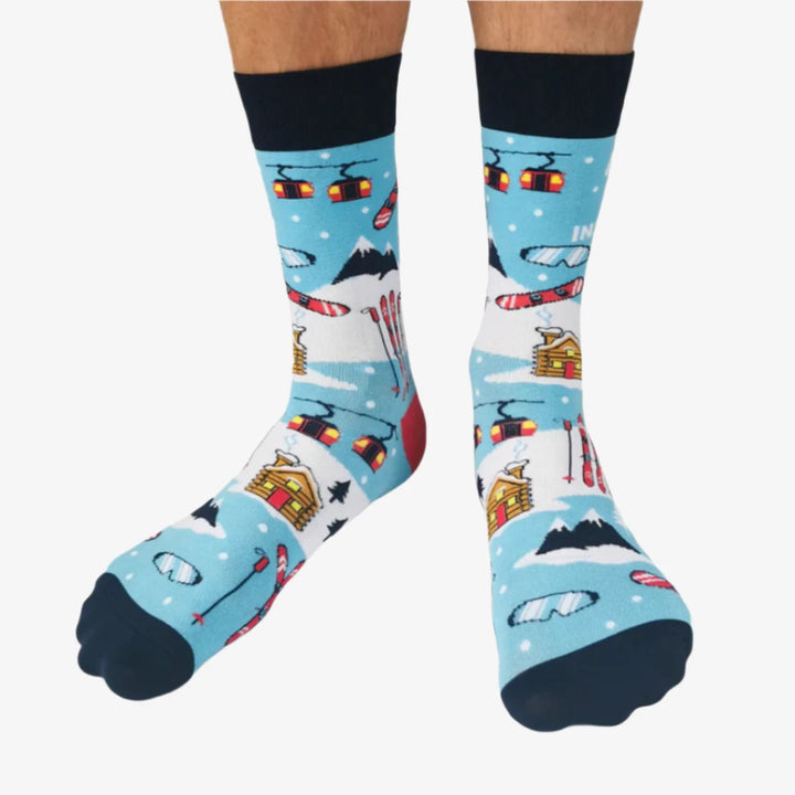 A pair of blue socks with a winter sports theme. The socks feature illustrations of skiers, snowboards, and a mountain cabin. The text 