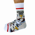 A pair of crew socks featuring a hockey theme. The socks are mostly grey with white and red stripes at the top and bottom. The design includes hockey-related icons like a trophy, skates, and a puck. The text "Hockey Dad" is printed on the socks.