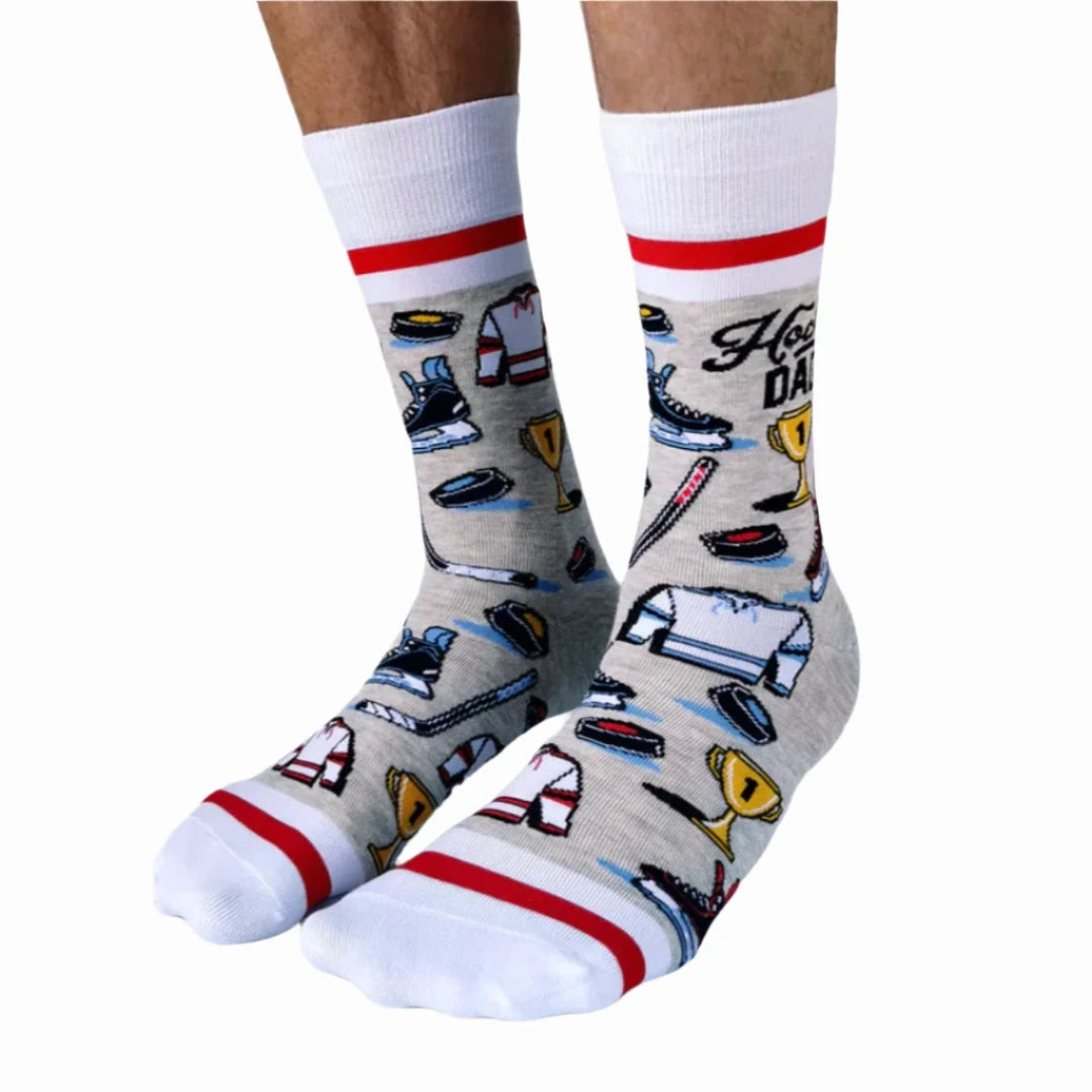 A pair of crew socks featuring a hockey theme. The socks are mostly grey with white and red stripes at the top and bottom. The design includes hockey-related icons like a trophy, skates, and a puck. The text "Hockey Dad" is printed on the socks.